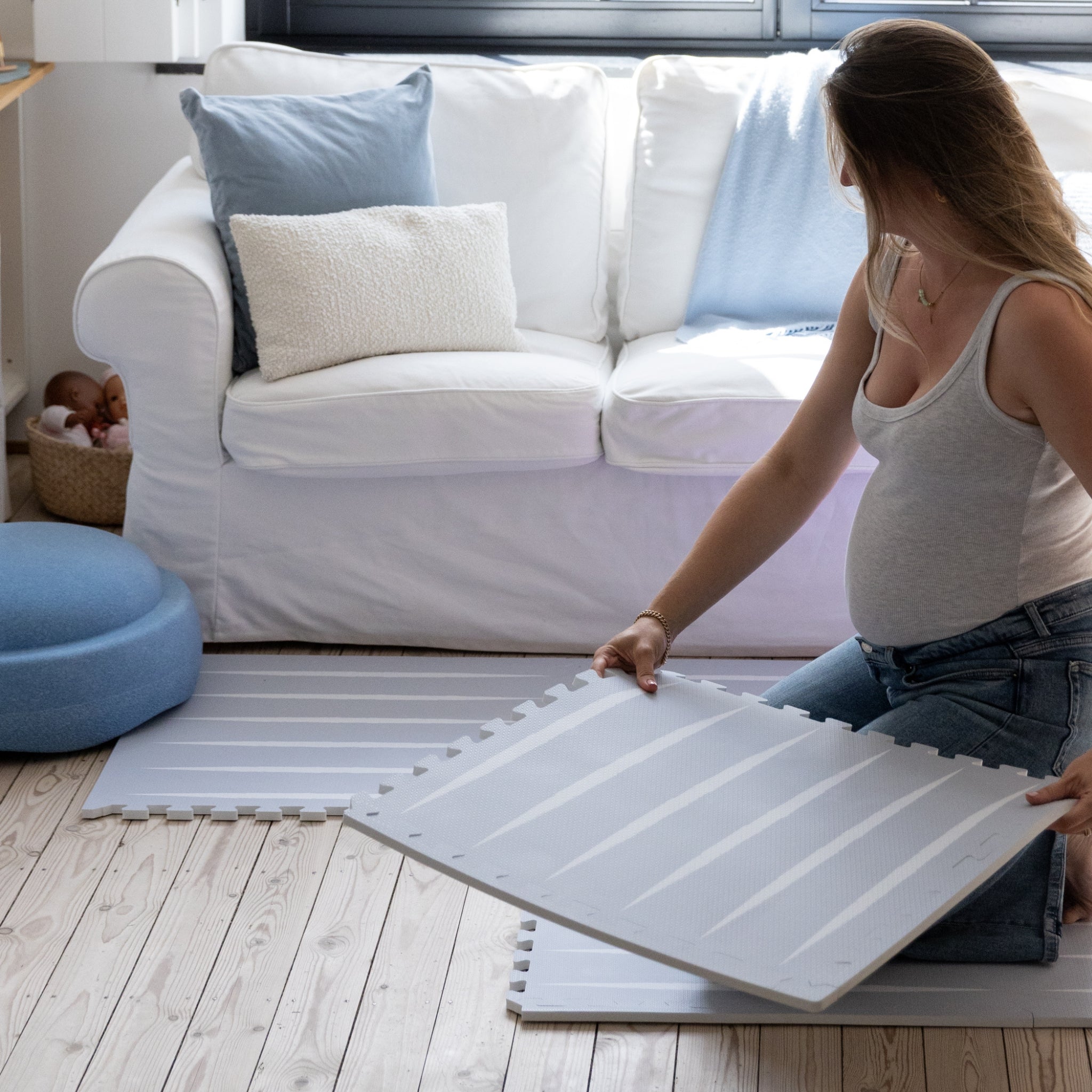 Zwangere vrouw klikt speelmat baby foam Lucio Nordic Mist van Eeveve in elkaar, met blauwe tinten die perfect matchen bij een witte sofa en houten vloer